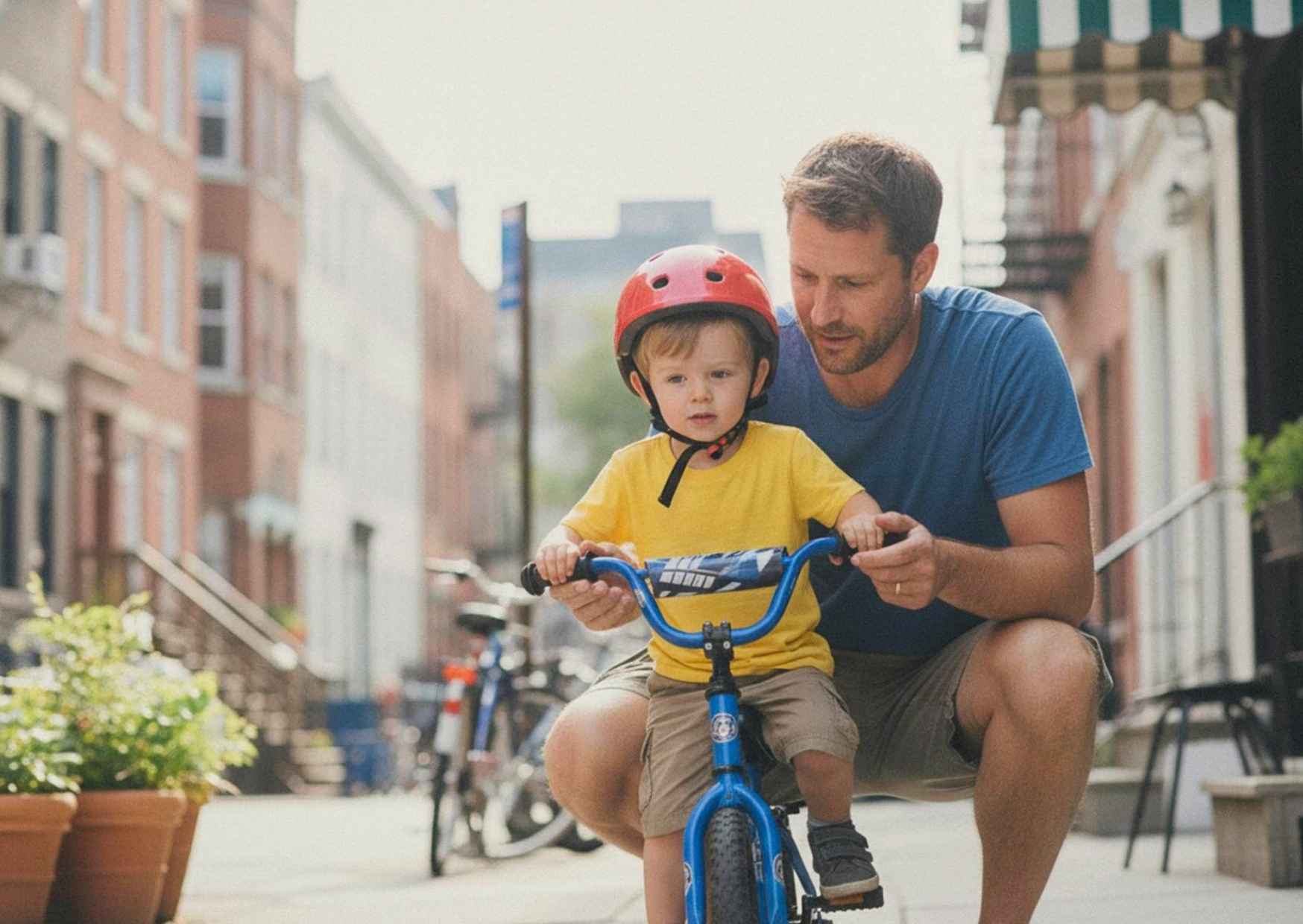 A man crouched on a sidewalk helping a child balance on a bike